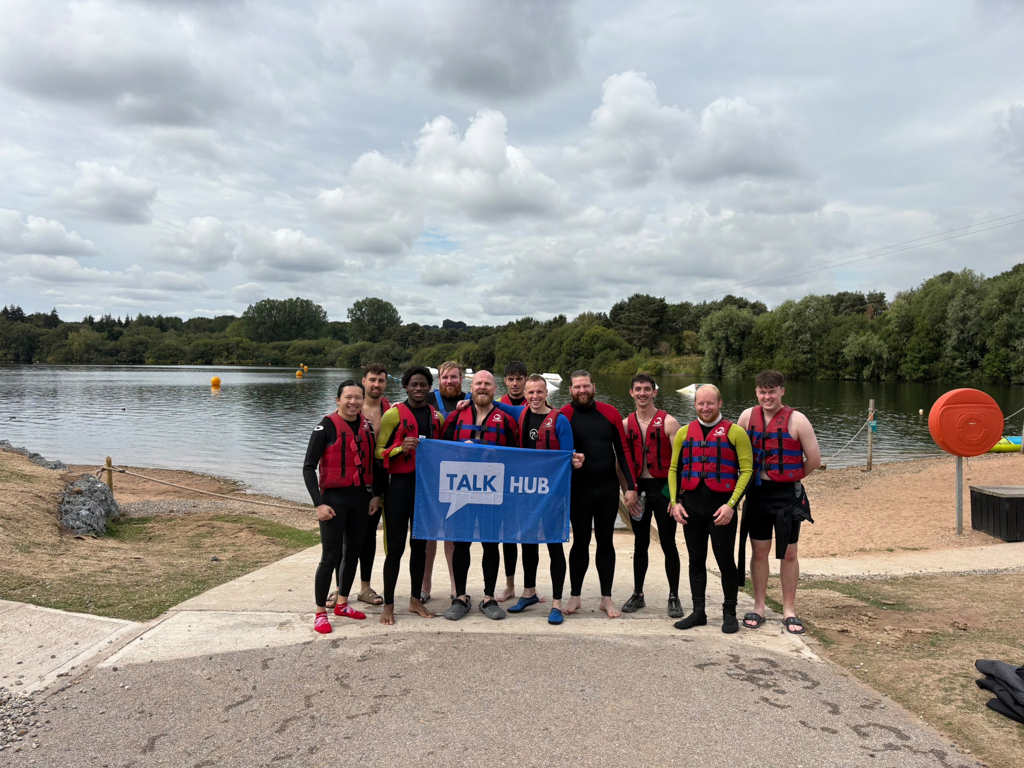 TalkHub lads in wetsuits and life jackets by a lake holding a TalkHub flag