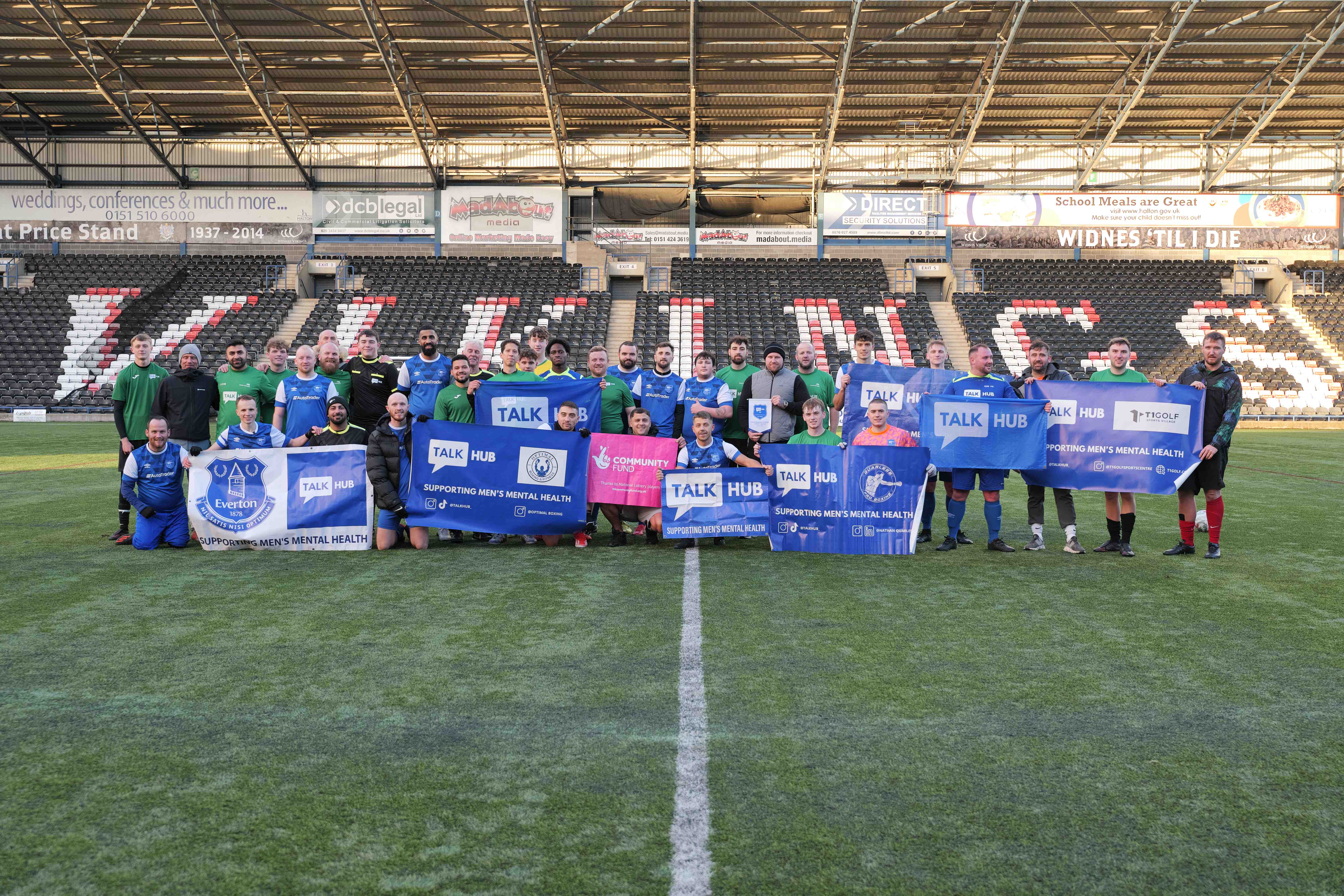 A large group of TalkHub men on the pitch at Widnes stadium holding banners
