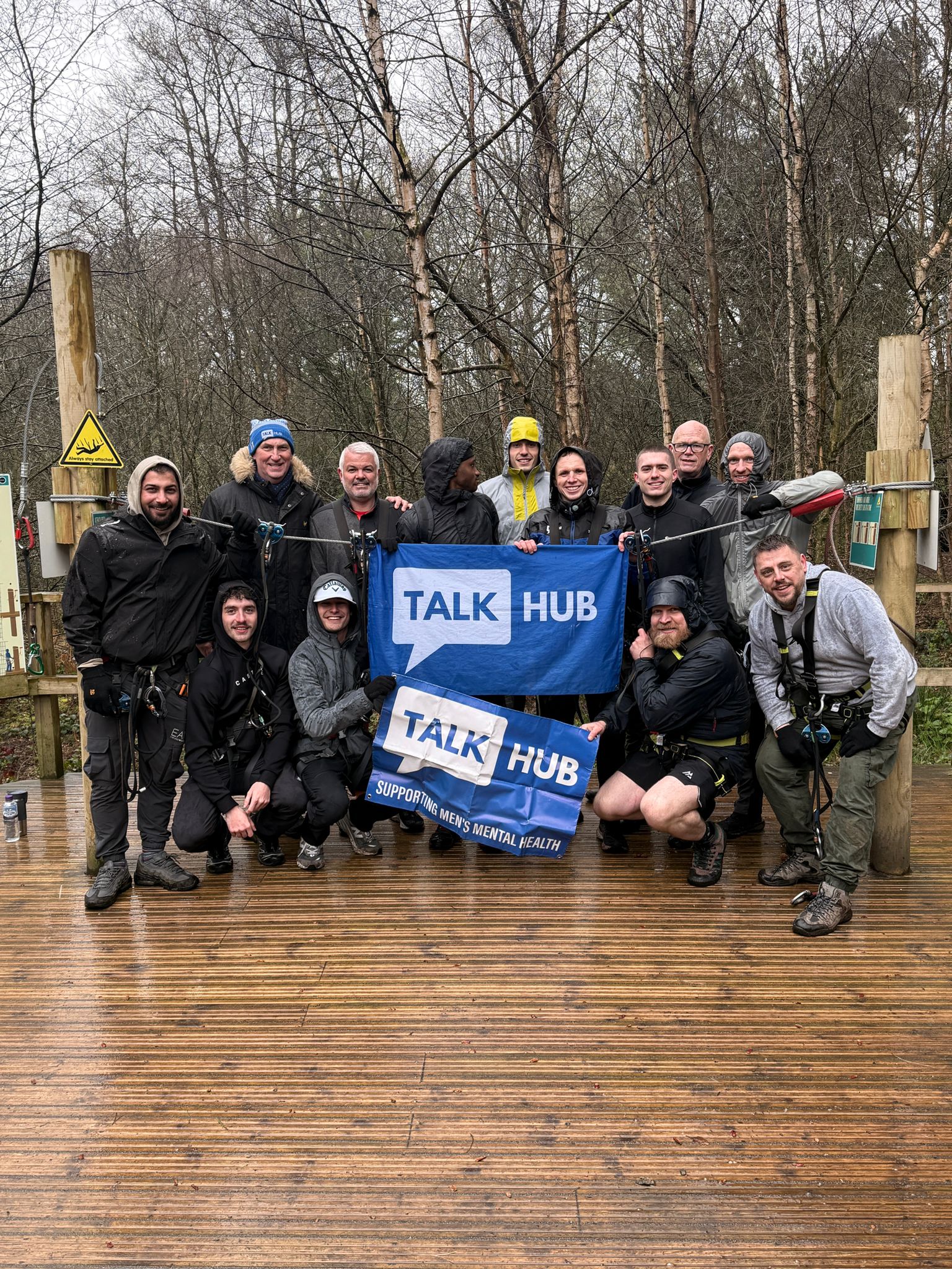 TalkHub group in climbing harnesses on a wet woodland platform at Go Ape holding TalkHub banners