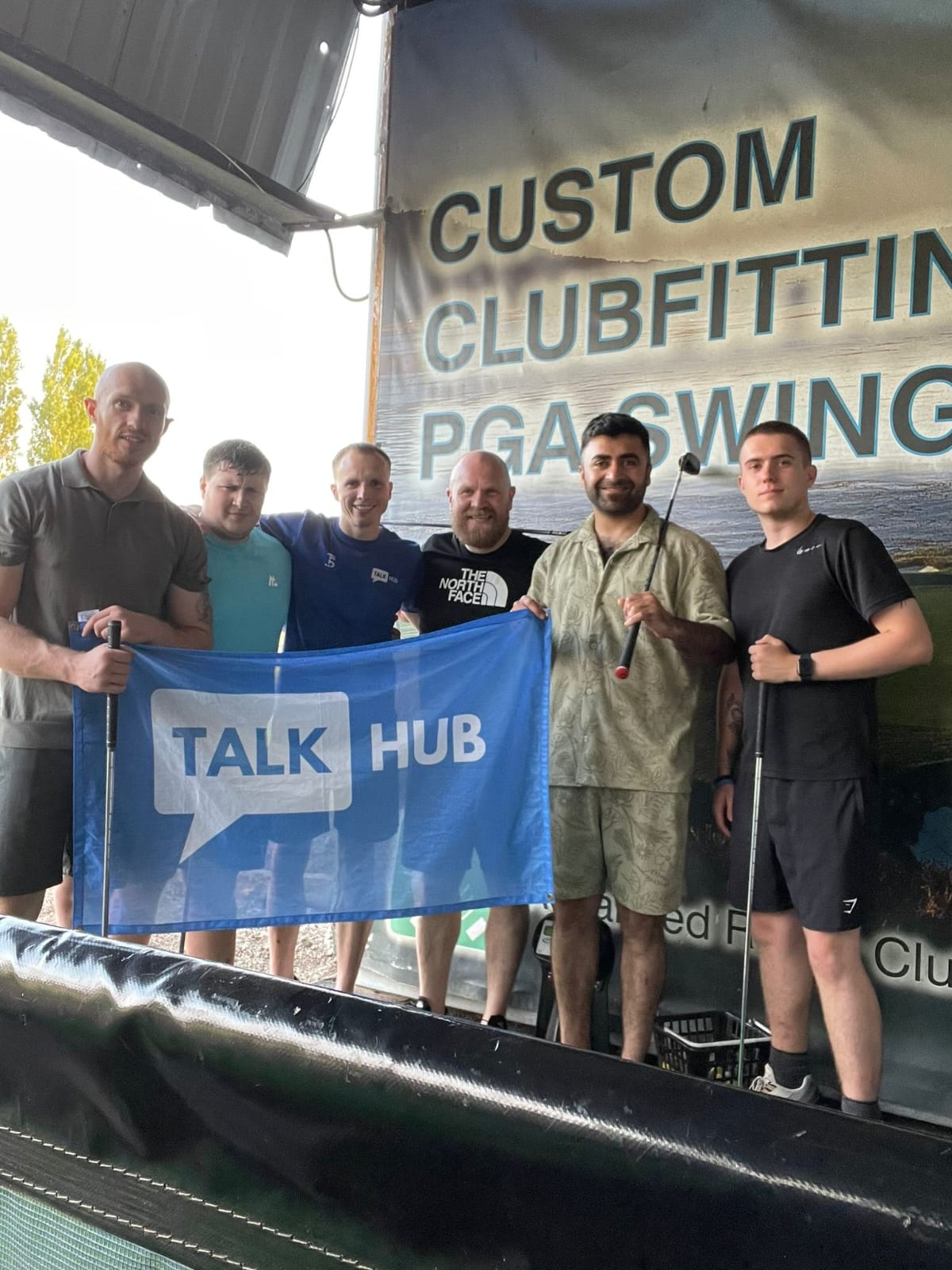 Six TalkHub members at a golf driving range holding golf clubs and a TalkHub flag