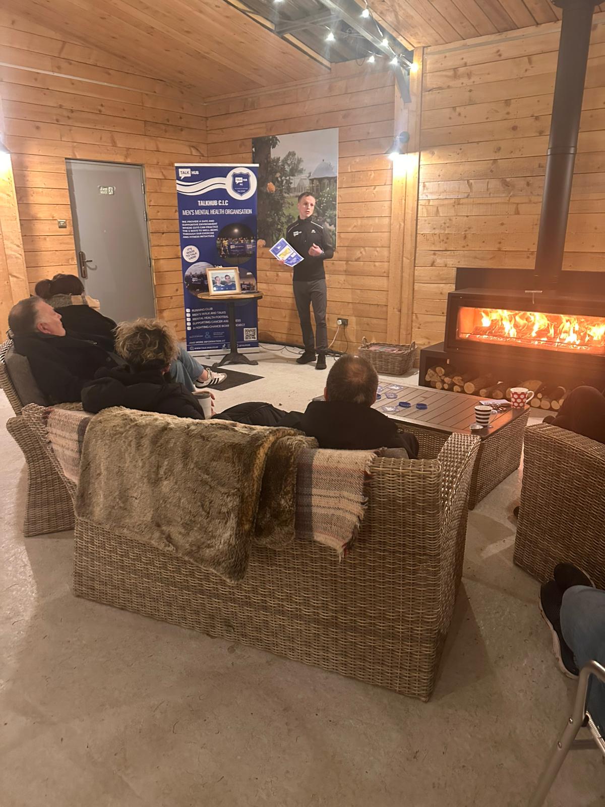 Jay giving a TalkHub talk to a small group seated on sofas beside a log burner