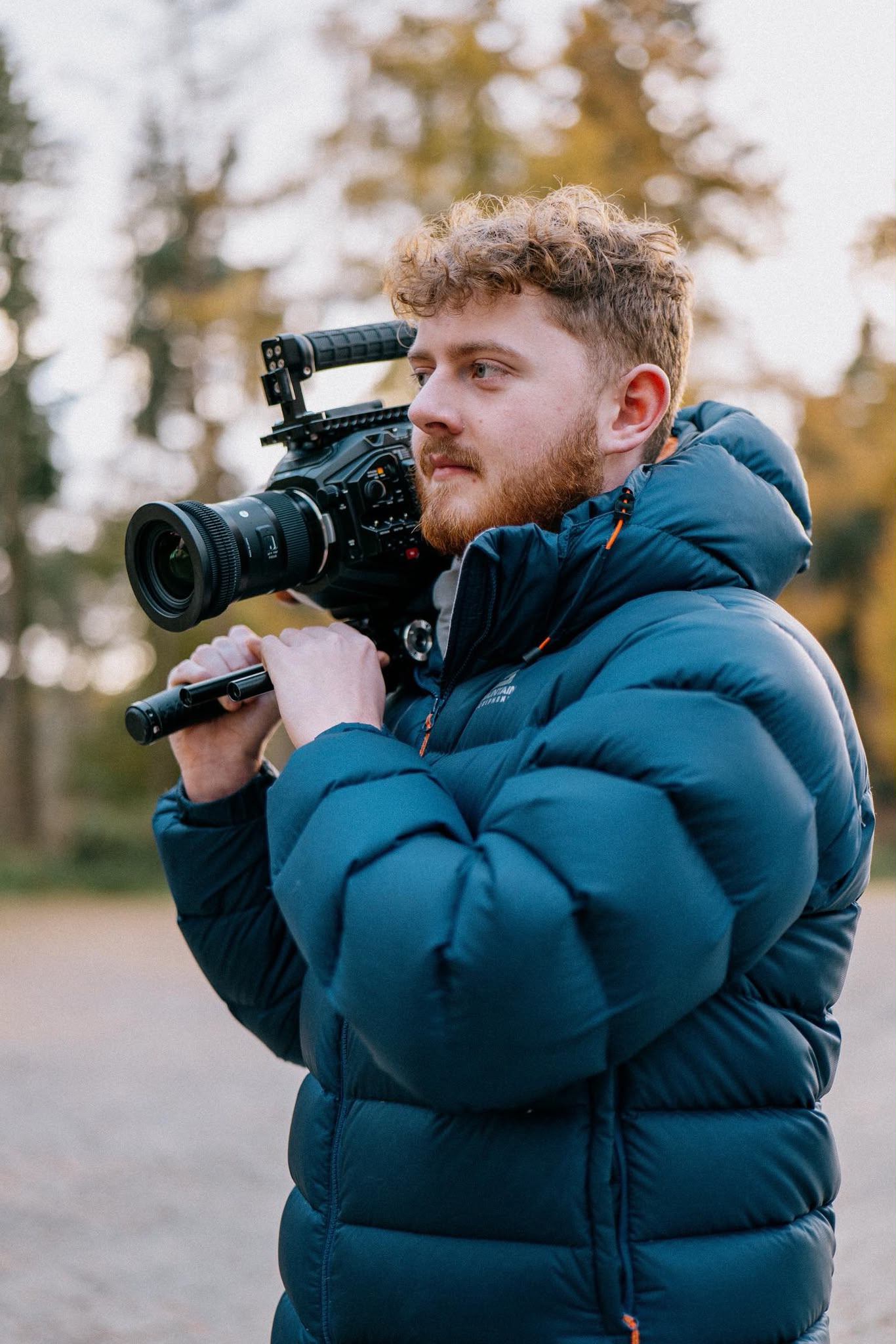 Aidan in a teal puffer jacket carrying a cinema camera on his shoulder in autumn woodland