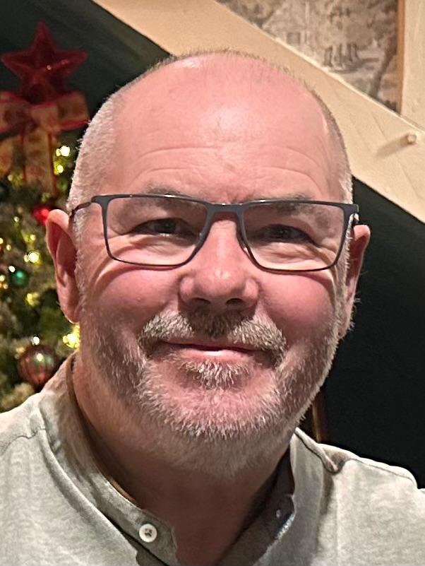 Tony smiling, wearing glasses and a light grey shirt, with a Christmas tree behind him